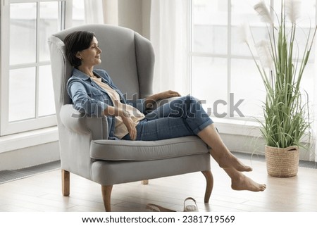 Serene attractive mature woman sit on armchair in modern living room looking away, enjoy lazy weekend, daydreaming, have peaceful leisure, relaxing alone at home. Lifestyle, cozy furniture, pastime Royalty-Free Stock Photo #2381719569