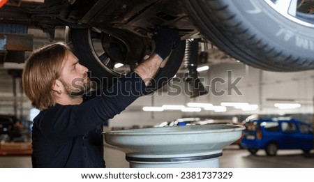 A male car mechanic in uniform changes the oil in the engine while standing under the car on the lift