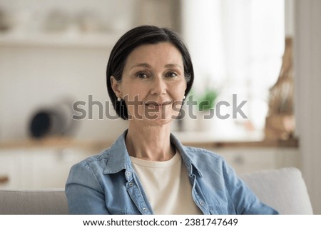Attractive middle-aged woman in casual shirt resting on cozy sofa smile look at camera spend pleasant leisure at modern own, cozy apartment. Carefree mature housewife, home-owner close up portrait