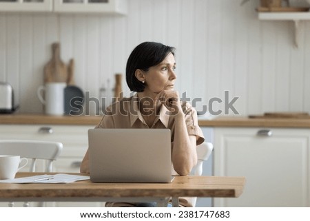Serious middle-aged woman sit at table in kitchen with laptop, working, managing family budget, doing freelance job, make online payments, staring into distance, ponder on problem, feels concerned