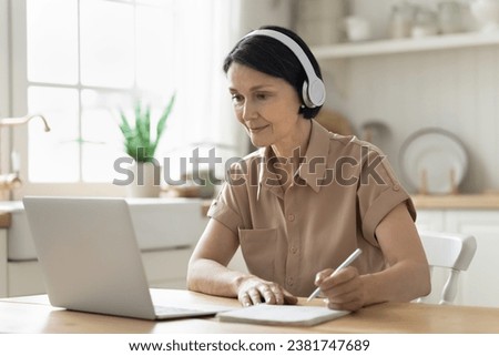 Middle-aged woman in headphones sit at table in kitchen look at laptop screen, makes notes, e-learns, listen audio lesson, gain new knowledge use modern technologies. Webinar, on-line class, education