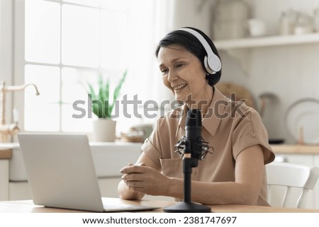 Aged woman talk into microphone sit at table in kitchen looks at laptop screen having remote talk, makes on-line communication with followers, take part in stream, recording webinar or podcast at home Royalty-Free Stock Photo #2381747697