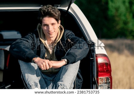 A young guy is resting lying in the open trunk of his car with a thermos in nature in a field