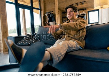 A young charismatic guy emotionally watches a film sitting on the couch and putting his feet up on table at home