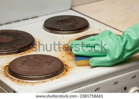 Woman hand in glove remaining burnt stains on white dirty electric stove scrubbing using sponge Royalty-Free Stock Photo #2381758661