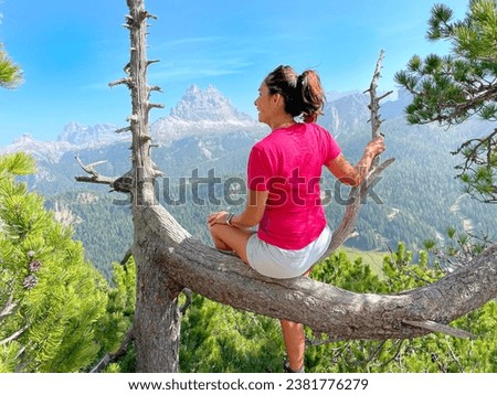 Sporting at an older age theme: sporty older woman sitting on a dry tree enjoying the view of the Dolomites panorama. Red shirt, shorts, dark hair, backpack, sunny day.  