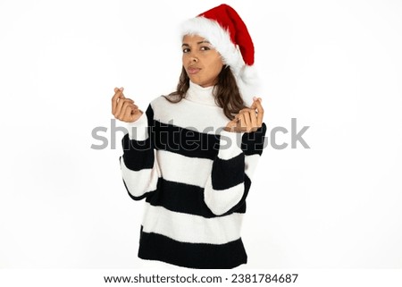 Young beautiful woman wearing striped sweater and a santa claus hat at christmas doing money gesture with hands, asking for salary payment, millionaire business