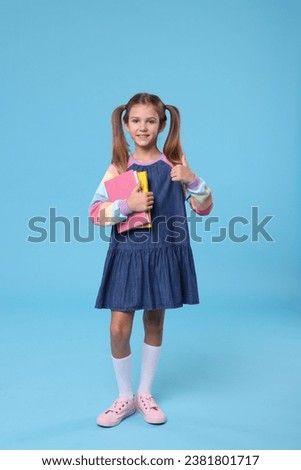 Happy schoolgirl with books showing thumb up on light blue background Royalty-Free Stock Photo #2381801717