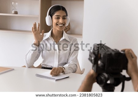 Happy young Indian business professional girl in headphones waving hand hello at camera. Cropped shot of photographer takin picture of happy model woman. Shooting backstage
