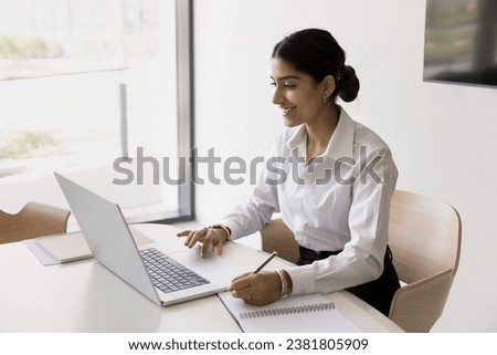 Happy young Indian office employee girl using laptop for Internet communication in office writing notes, smiling, talking on video call. Company intern watching business webinar Royalty-Free Stock Photo #2381805909