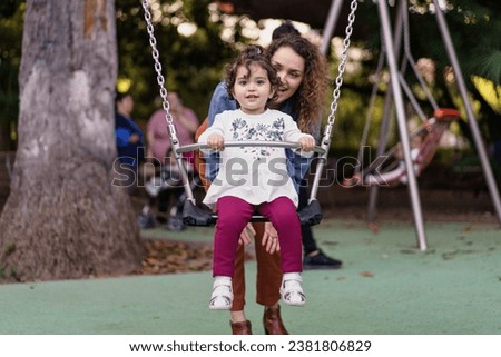 A heartwarming image of a young girl enjoying her time on a swing, with the presence of her mother right behind her. Their smiles and the playful atmosphere highlight the moments families share. Royalty-Free Stock Photo #2381806829