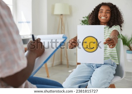 Happy little African American school child girl sitting on chair and showing picture with yellow smiling emoji while her school psychologist or therapist is taking notes. Psychology, emotions concept