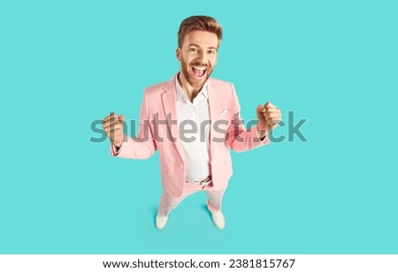 Happy guy celebrating his success and having fun. High angle shot of joyful young man in pink suit standing on blue color background, looking at camera, smiling and doing YES gesture. Fashion concept Royalty-Free Stock Photo #2381815767