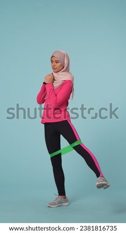 Full-sized isolated photo capturing an attractive young woman wearing a sportswear and a hijab, sheila. She is exercising with a green rubber exercise band.