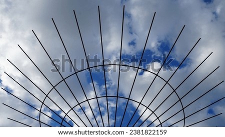 A metal structure with thin rods forming a semi-circle against a blue sky with white clouds. Royalty-Free Stock Photo #2381822159