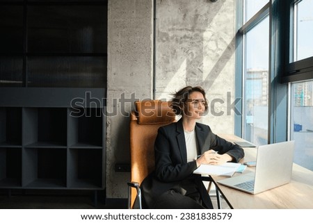 Portrait of successful lady boss, businesswoman in an office, working on her laptop, looking outside window, signing documents and papers. Royalty-Free Stock Photo #2381838579