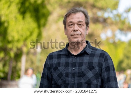 Street portrait of a relaxed 40-50 year old man wearing a shirt against a neutral blurred background of a city park. Perhaps he is a shopper, actor or lorry driver, military pensioner. Royalty-Free Stock Photo #2381841275