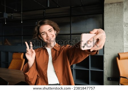 Portrait of young woman, social media content creator in office, posing for selfie, taking pictures on smartphone, posing for photo on mobile phone.