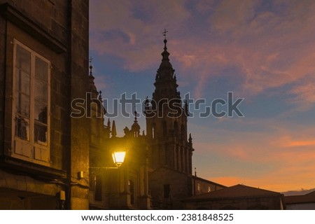 In the foreground, a lit lantern, in the background, stands one of the towers of the Cathedral of Santiago de Compostela, with a beautiful sunset sky in the background