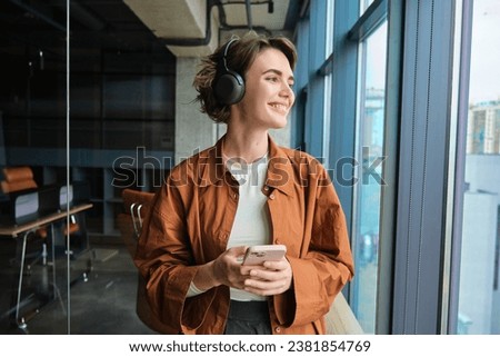 Portrait of woman working in office, digital nomad in casual clothes, wearing wireless headphones, holding mobile phone, looking outside window with pleased smile. Royalty-Free Stock Photo #2381854769