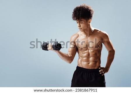 Athletic muscular young brazilian man lifting dumbbell, flexing arms muscles while training over gray studio background, posing shirtless. Fitness and strength concept. Free space