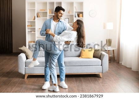 Young Indian couple, in casual attire, shares playful dance move in their homey living room, filled with natural light, contemporary furniture, full length, free space Royalty-Free Stock Photo #2381867929