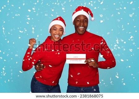 Happy black man and woman holding shopping gift certificate card and gesturing yes, posing in santa hats expressing joyful emotions on blue studio backdrop with falling confetti
