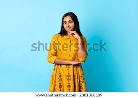 Pretty Indian young woman in yellow traditional dress posing touching face, smiling to camera while standing over blue studio background. Banner for beauty and fashion advertisement Royalty-Free Stock Photo #2381868189