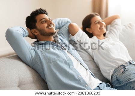 Happy Hindu spouses seated on sofa, placing hands relaxed behind their heads, exchanging delighted smiles in leisurely, homey ambiance Royalty-Free Stock Photo #2381868203