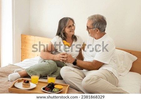 Glad senior european man give flower to lady on bed, have romantic breakfast, enjoy good morning in light bedroom interior. Rest at weekend together, relationships and love