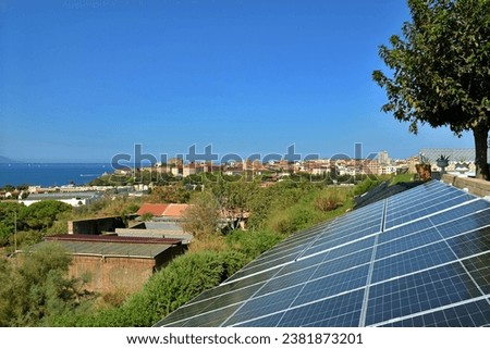 The sun's rays shine brightly on solar panels installed on a hillside, against the backdrop of the city and the sea. Example of installation and use of solar panels. Green energy concept. Copy-space. Royalty-Free Stock Photo #2381873201