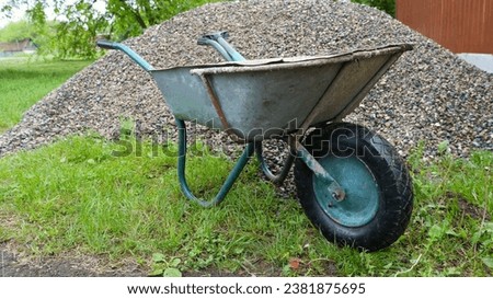 old garden cart standing next to a pile of gray gravel on a green lawn in a rural area, manual transportation of crushed stone on a wheelbarrow for use in a private household Royalty-Free Stock Photo #2381875695