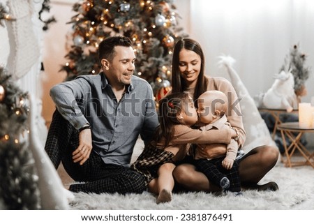 Happy family having fun against the background of a New Year tree and Christmas decor. Young dad and happy mother with son and daughter smile and enjoy New Year holidays. Sister kissed brother