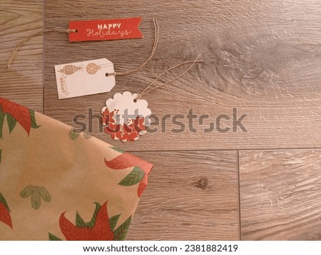 Three christmas gift tags and some rustic wrapping paper with the pictures of poinsettias on the wooden table.
