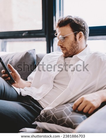 Concentrated millennial man searching phone number on mobile resting at home interior,