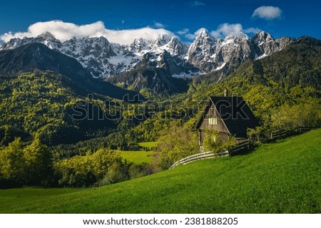 Amazing spring alpine scenery with cute wooden hut on the green field and high snowy mountains in background, Srednji Vrh village, Kranjska Gora, Julian Alps, Slovenia, Europe Royalty-Free Stock Photo #2381888205