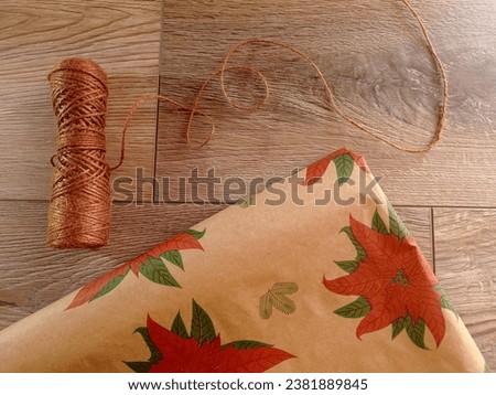 Rustic christmas gift wrapping materials. A ball of jute string and some wrapping paper with the pictures of poinsettias on the wooden table.