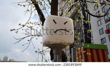 Lamppost with a glass white cube depicting a sad smiley face against the background of the city surroundings
