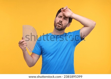 Unhappy man with hand fan suffering from heat on orange background