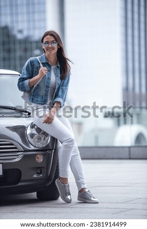 Full length image of beautiful girl in jean jacket and sun glasses looking at camera and smiling while leaning on her car