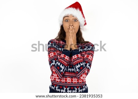 Young beautiful woman wearing knitted sweater and a santa claus hat at christmas keeps hands on mouth, looks with eyes full of disbelief, being puzzled with amount of work