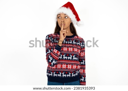 Young beautiful woman wearing knitted sweater and a santa claus hat at christmas silence gesture keeps index finger to lips makes hush sign. Asks not to share secret