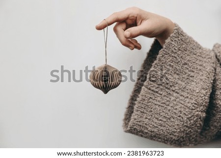 Merry Christmas still life. Female, child hand holding paper christmas ornament on white background. Preparation for winter holidays, decorating christmas tree with stylish bauble. Beige sweater. 