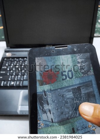focus on the display of Malaysia money on the smartphones screen. there is a hand holding it and there is a laptop in the background
