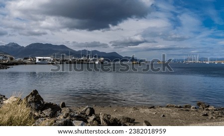 In the gulf of the sea, in the port of Ushuaia, there are ships, yachts, boats with masts. Andes mountains on a background of blue sky, clouds. In the foreground is a rocky shore. Argentina. 
