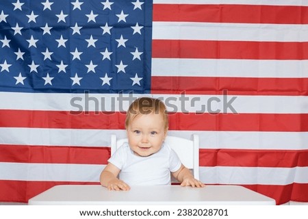 Positive toddler girl sits at white table on background of USA flag. Concept of nationality and patriotism in childhood. Girl moves desk sitting on chair