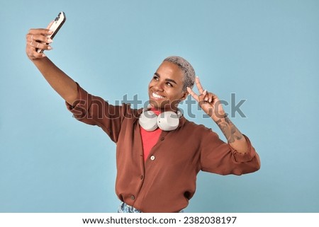 Young cheerful pretty African American woman takes selfie on mobile phone and demonstrates victory gesture wanting to become popular blogger stands on plain turquoise background. Internet celebrity
