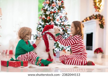 Christmas at home. Kids and dog under Xmas tree. Little boy and girl hug pet in Santa hat and open Christmas presents. Children play with animal. Winter holiday celebration. Love and friendship.