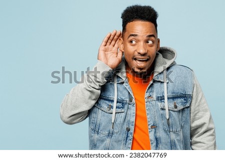 Young curious nosy man of African American ethnicity wearing denim jacket orange t-shirt try to hear you overhear listening intently isolated on plain pastel light blue background. Lifestyle concept Royalty-Free Stock Photo #2382047769