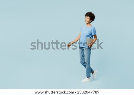 Full body side profile view smiling happy young IT woman of African American ethnicity wear t-shirt casual clothes hold closed laptop pc computer isolated on plain pastel light blue cyan background Royalty-Free Stock Photo #2382047789